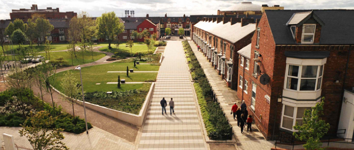 A photo from a high position showing a shared green space by a row of victorian terraced houses with large concrete paving boulevards that people are walking along.