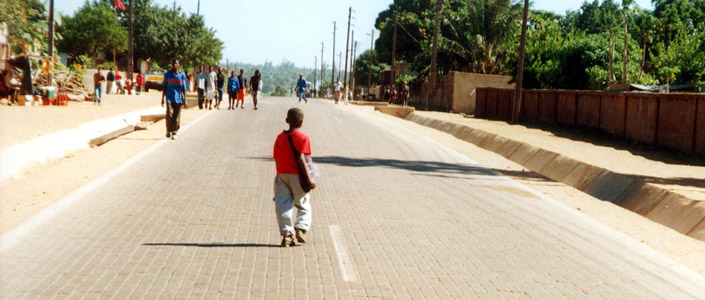 A photo of a concrete paving block road with a child walking along it in the foreground and groups of people further along in the distance.