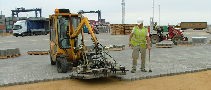 A mechanical concrete block laying machine in use on a work site, there is a man in high-vis operating it, and another man to the right also in high-vis monitoring the alignment of the blocks being layed.